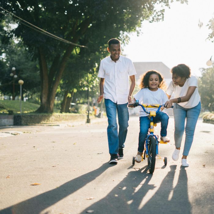 Happy Family Teaching Their Child To Cycle On A Sunny Day Outdoors.