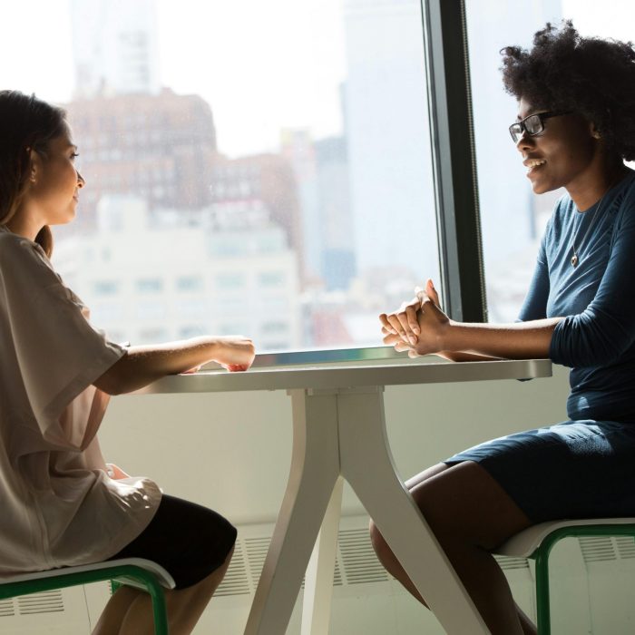 Two Women Sitting And Talking At A Table With A City View From The Window.