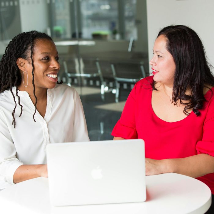 Two Women In An Office Discussing Work Using A Laptop, Smiling And Engaged.