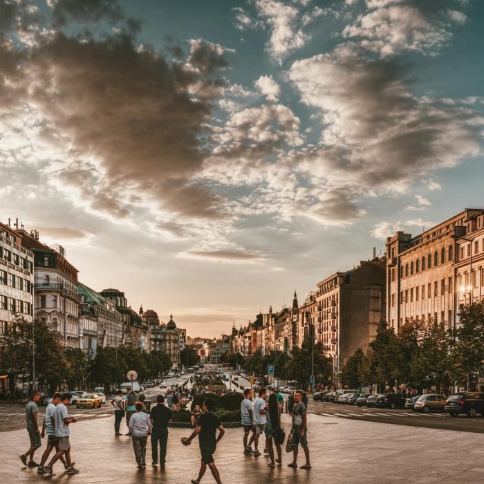 A Lively City Square At Sunset With People Walking, Historic Architecture, And Dramatic Cloudy Sky.