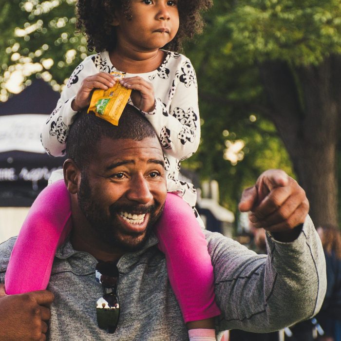 A Joyful African American Father With Curly Haired Daughter On Shoulders, Outdoors.
