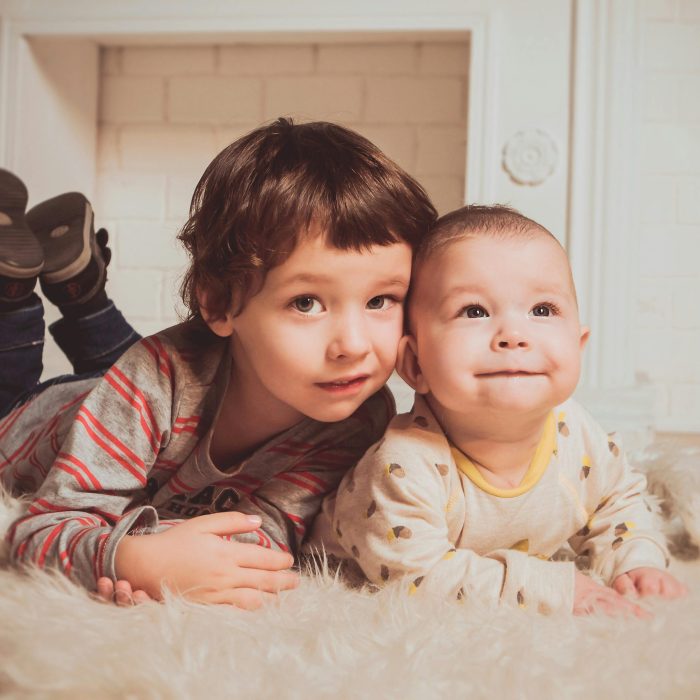 Two Young Siblings Lying On A Fluffy Rug Indoors, Exemplifying Warmth And Family Bonding.