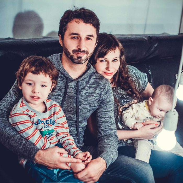 A Joyful Family Of Four Sitting On A Sofa, Smiling Warmly Indoors.