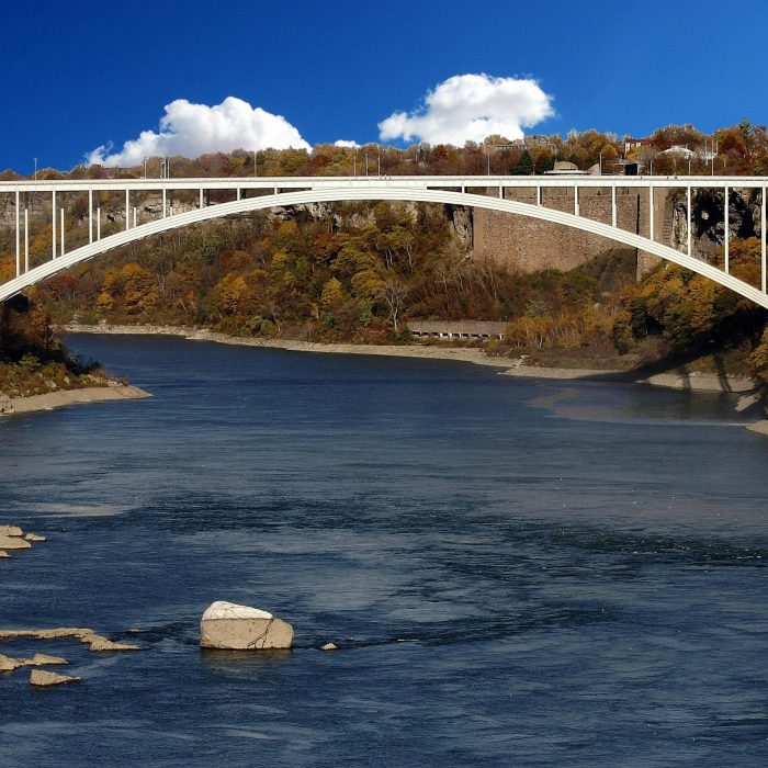 Panoramic View Of Rainbow Bridge Over The River In Fall With Vibrant Foliage And Blue Sky.