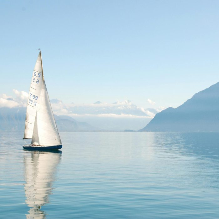 Sailing Boat Glides Peacefully On A Misty Lake Surrounded By Mountains, Under Clear Daylight.