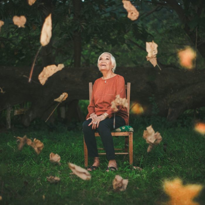 An Elderly Woman Sits Outdoors Enjoying The Falling Autumn Leaves, Capturing A Serene Moment.