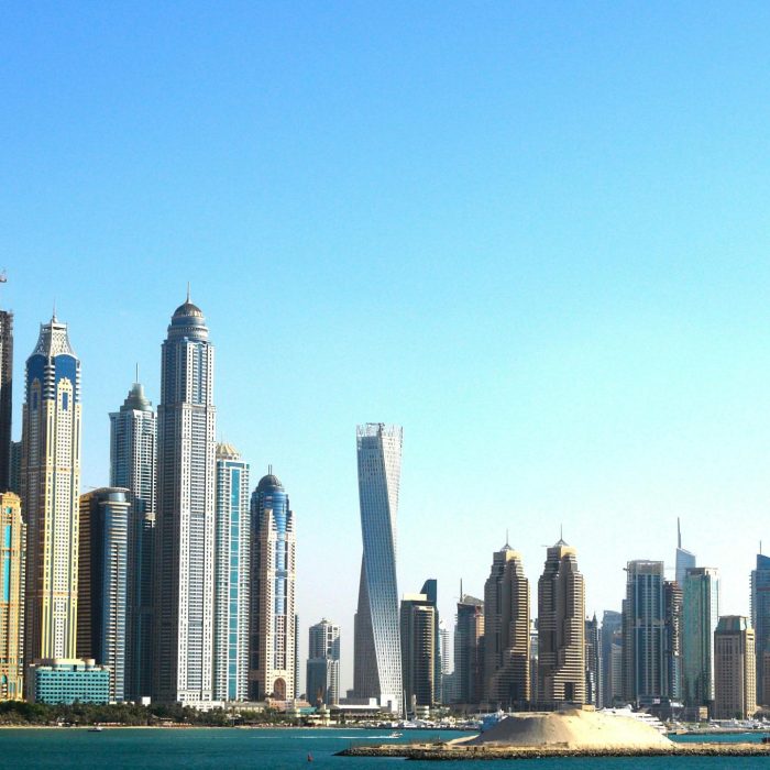 Panoramic View Of Dubai's Towering Skyscrapers Set Against A Vibrant Blue Sky And Waterfront.