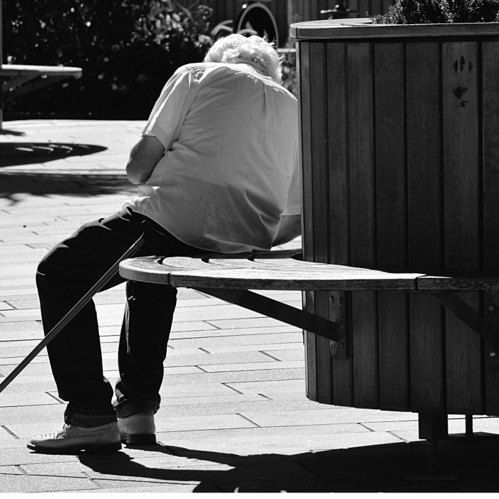 A Black And White Image Of A Senior Man Sitting On A Bench With A Cane In An Urban Park.