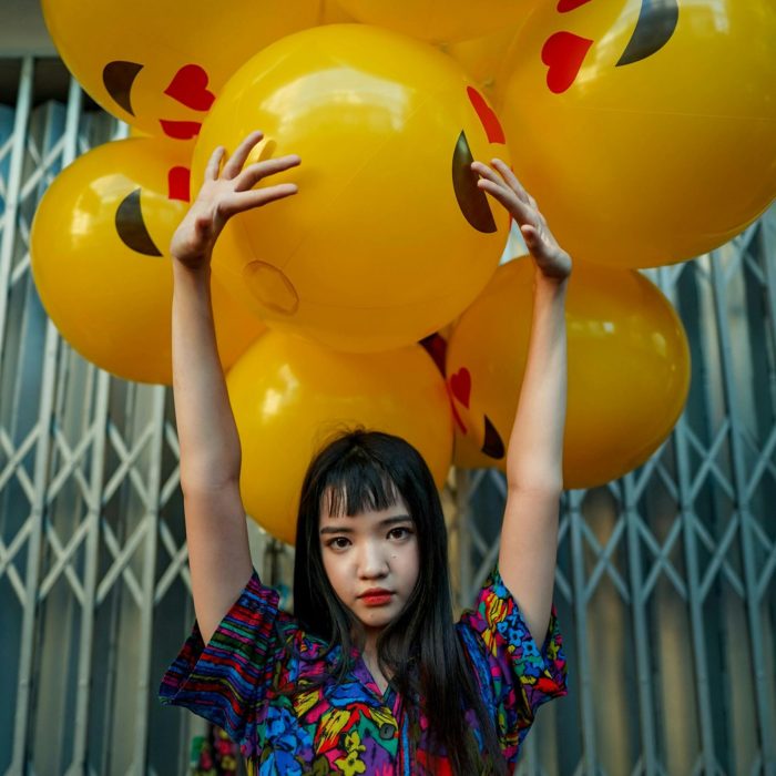 A Young Woman Poses With Bright Emoji Balloons Against An Urban Background.