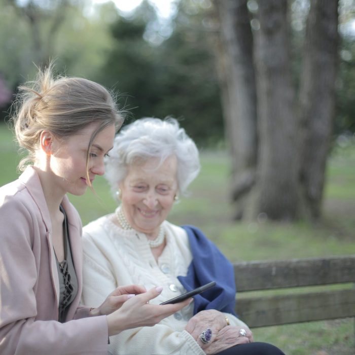 Side View Of Smiling Adult Female Helping Aged Mom In Using Of Mobile Phone While Sitting Together In Park