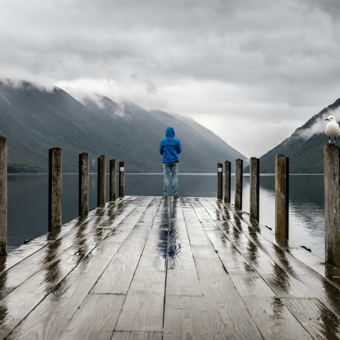 Person In Blue Jacket Stands On A Rainy Dock Overlooking Serene Lake And Mountains.