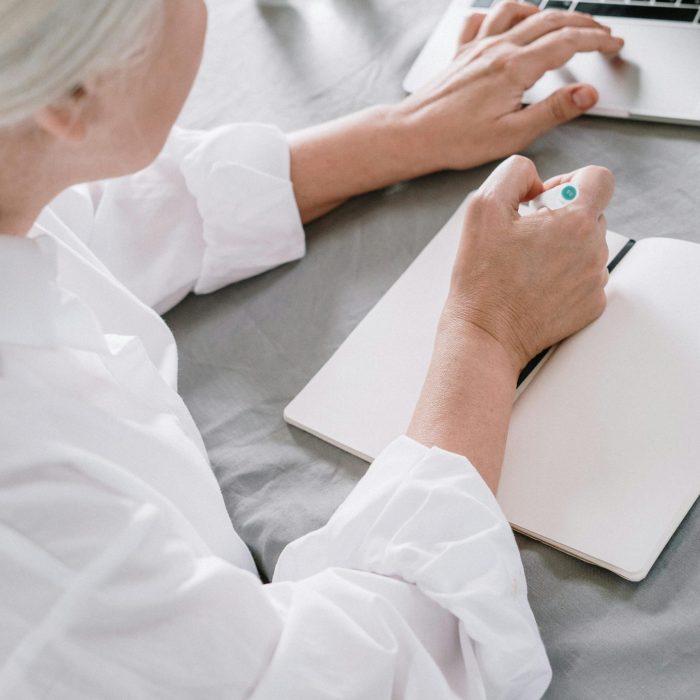 Elderly Woman Takes Notes While Using A Laptop At Home.