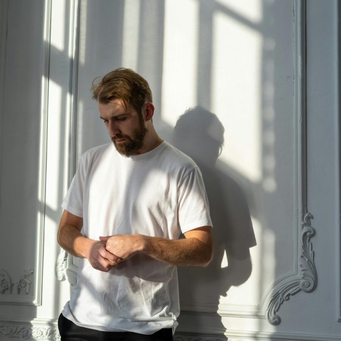 A Thoughtful Bearded Man In A White T Shirt Stands In A Sunlit Room With Ornate Walls.