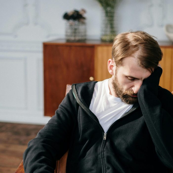 Bearded Man In A Black Hoodie Sitting In A Chair Indoors, Appearing Deep In Thought.