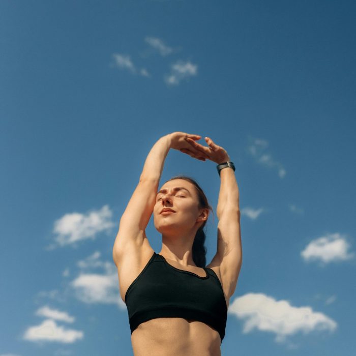 A Woman In Black Leggings And Sports Bra Stretches Under A Clear Blue Sky, Promoting Fitness And Freedom.