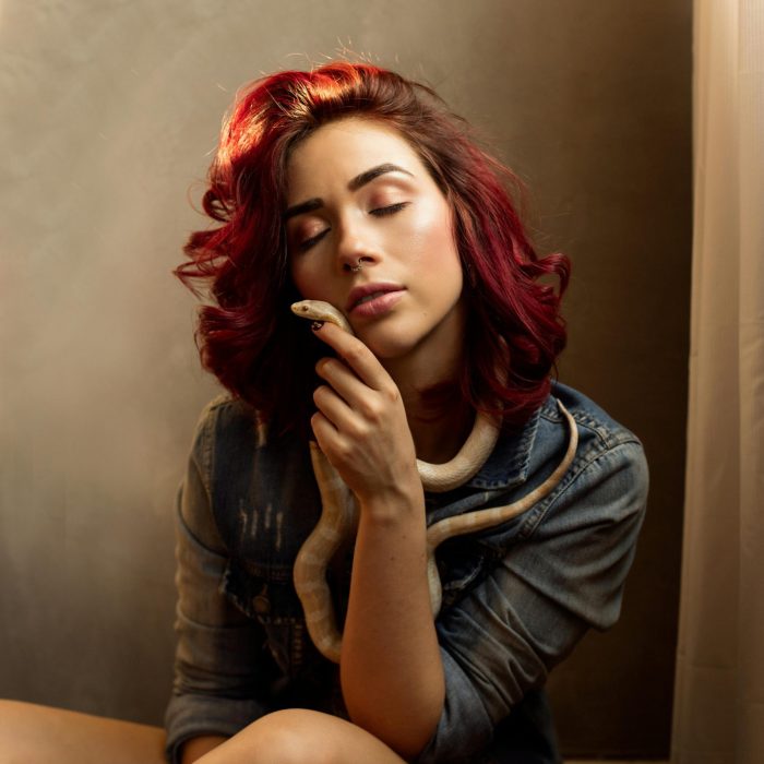 A Woman With Red Hair Holding A Snake, Eyes Closed, Wearing A Denim Jacket Indoors.