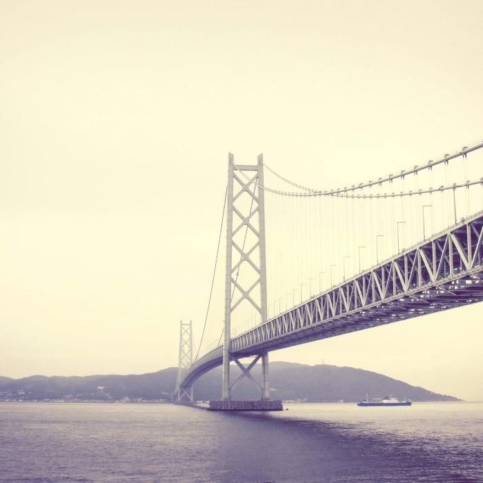 A Stunning View Of A Suspension Bridge Spanning The Sea With A Hazy Backdrop.