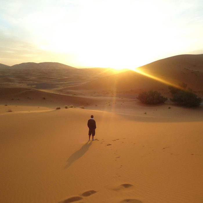 Silhouette Of A Man Walking Along Sand Dunes During A Sunrise In The Sahara Desert.