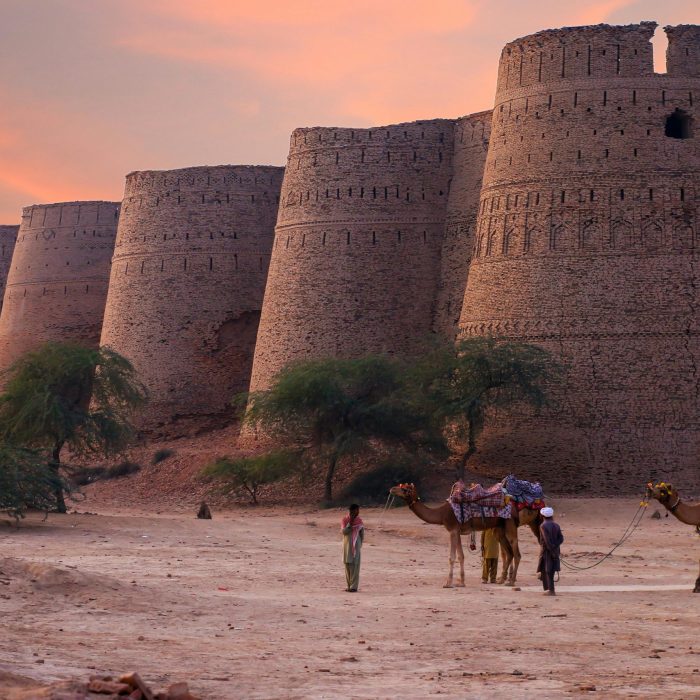Scenic View Of Derawar Fort's Towers At Sunset In The Punjab Desert With Camels And People.