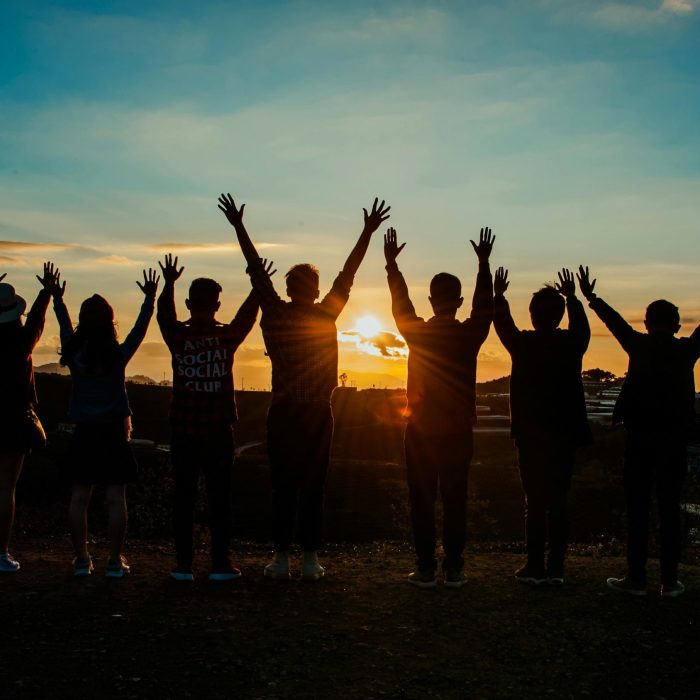 A Diverse Group Of Friends Raises Their Arms In Celebration Against A Vibrant Sunset Backdrop.