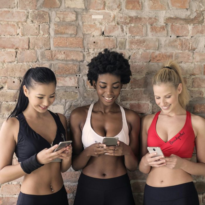 Three Diverse Women Smiling And Using Smartphones Indoors Against A Brick Wall.