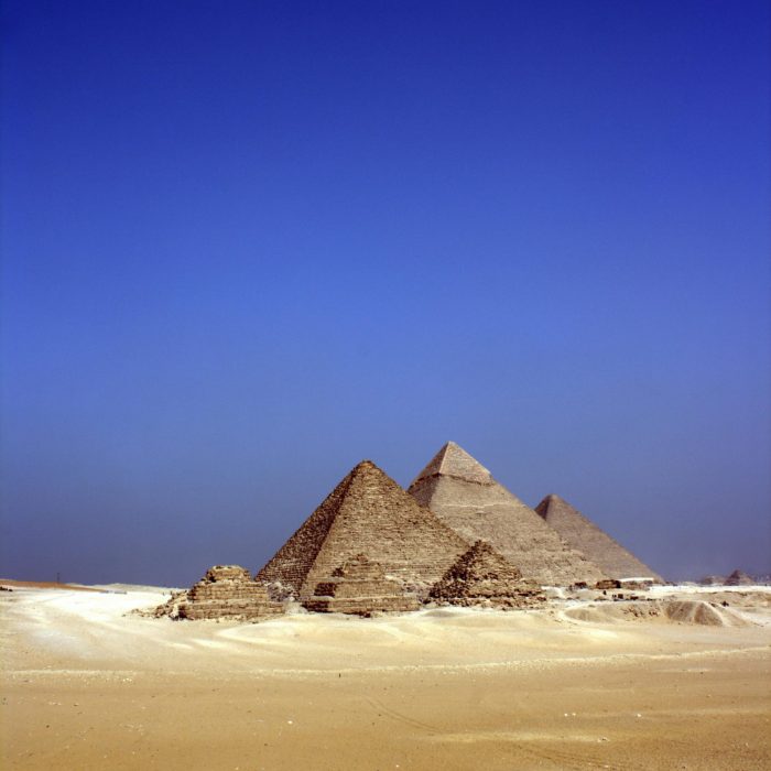 Stunning View Of The Pyramids Of Giza With A Lone Figure And Camel Under A Clear Blue Sky.