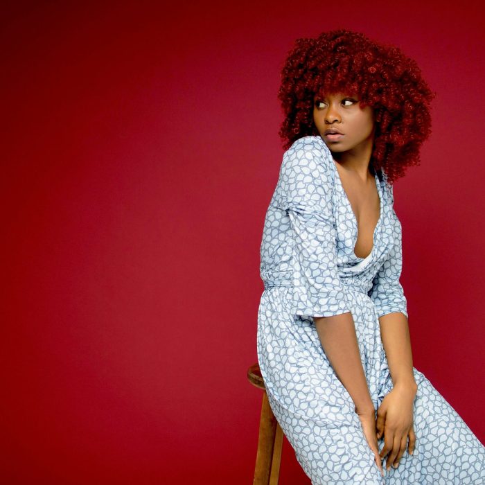 Portrait Of A Young Woman With Red Hair Sitting On A Stool Against A Red Backdrop, Showcasing Modern Fashion.